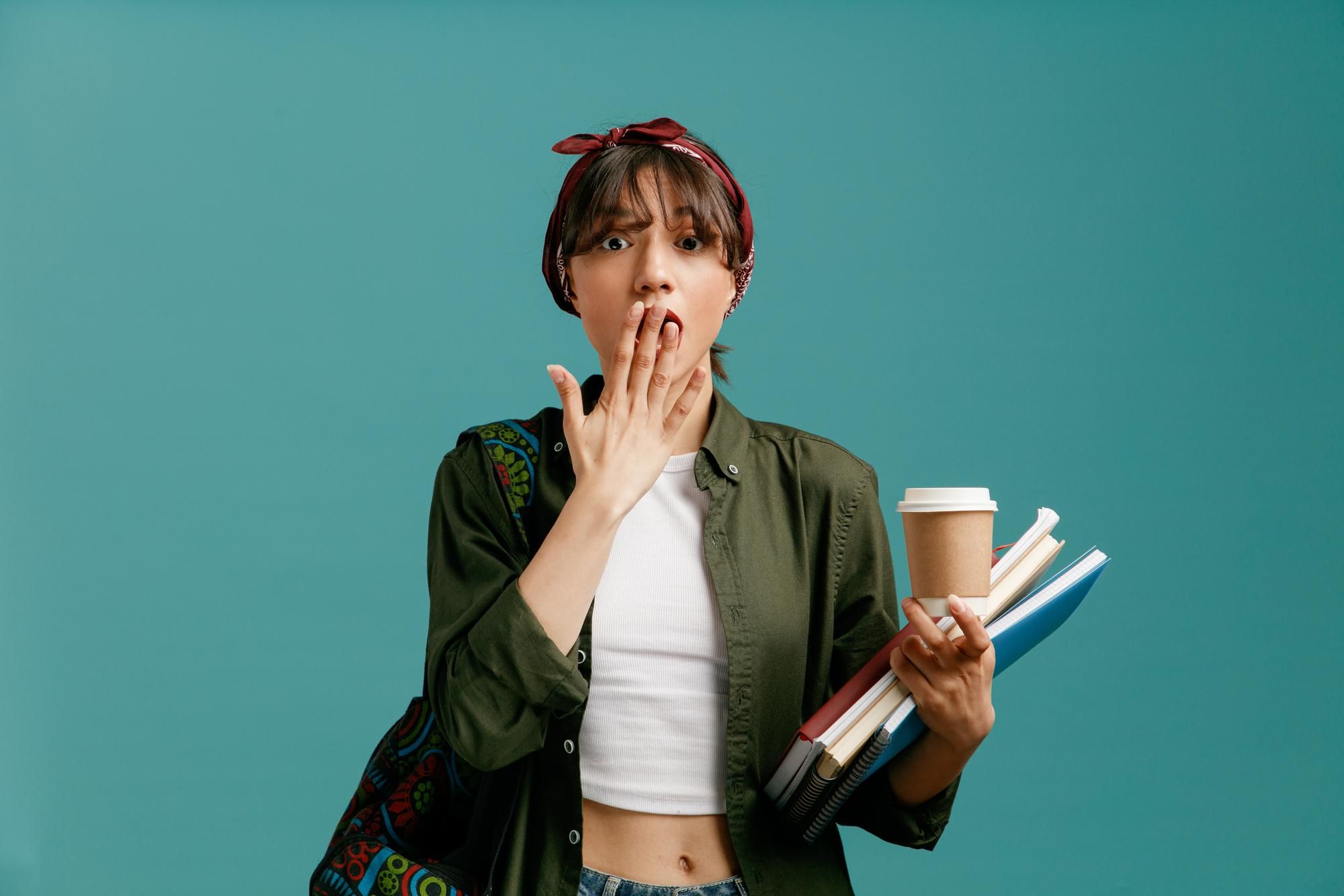 surprised-young-student-girl-wearing-bandana-backpack-holding-paper-coffee-cup-note-pads-looking-camera-making-oops-gesture-isolated-blue-background.jpg
