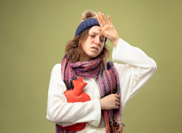 weak-young-ill-girl-wearing-white-robe-winter-hat-with-scarf-holding-hot-water-bag-putting-hand-forehead-isolated-olive-green_141793-86416.jpg